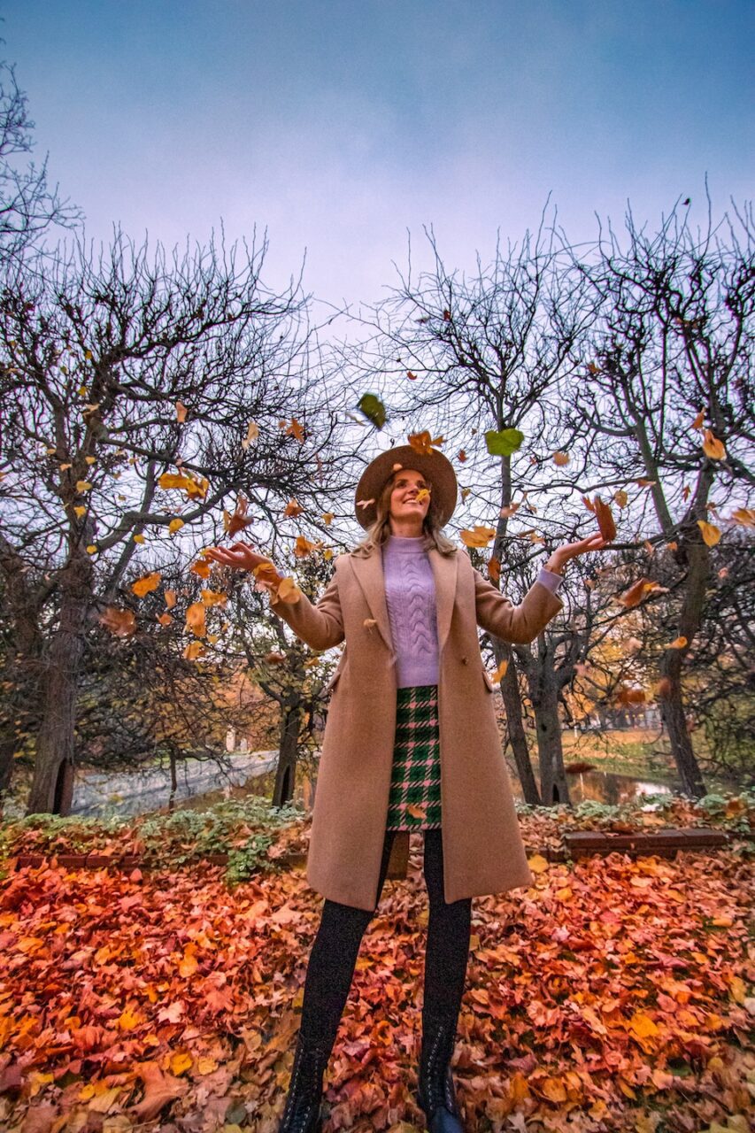 Kobieta w beżowym płaszczu i kapeluszu rzuca liście w parku jesienią w Polsce