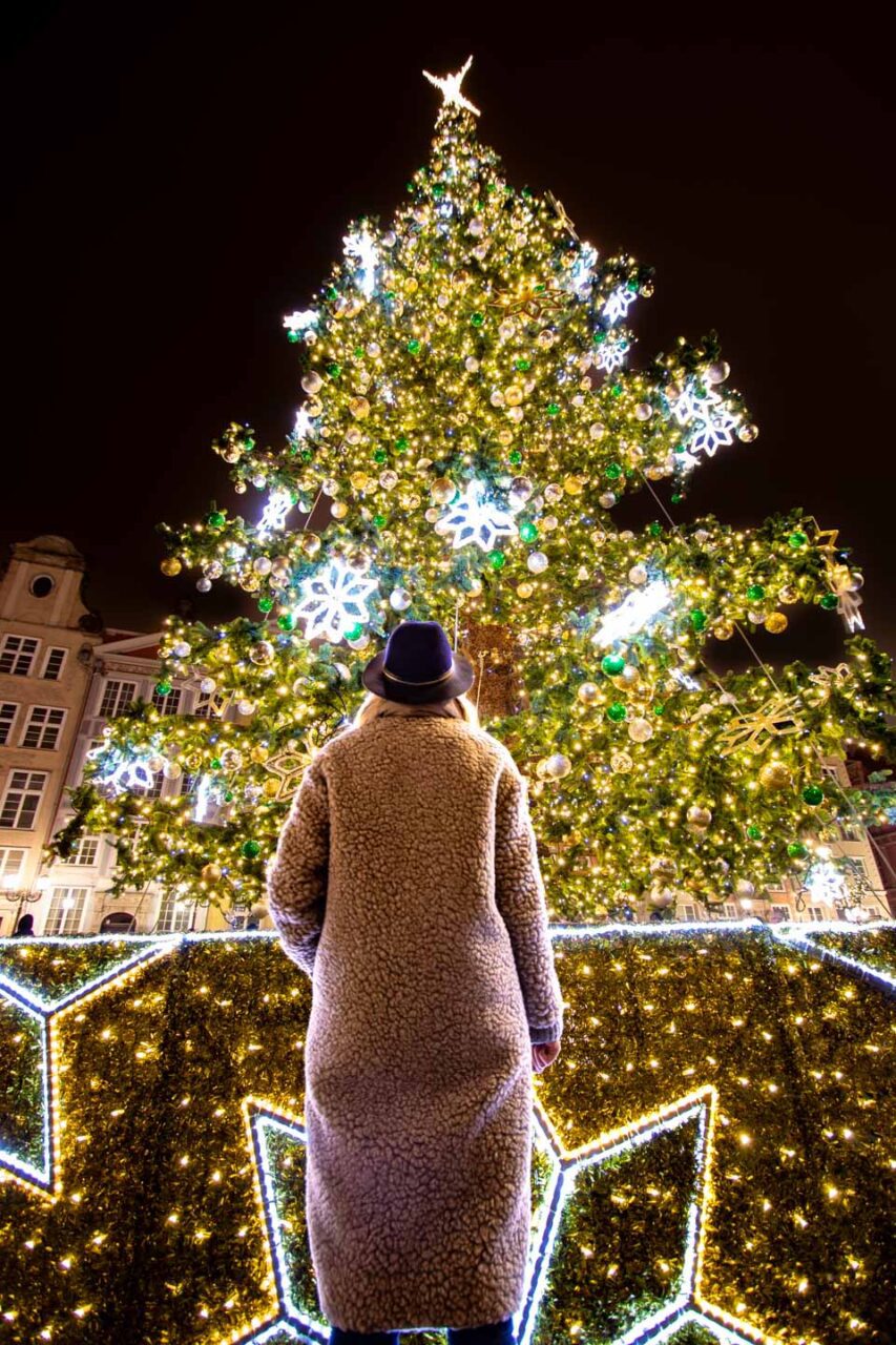 A woman in a teddy coat and hat looks at a huge, illuminated Christmas tree at a Christmas market in Europe