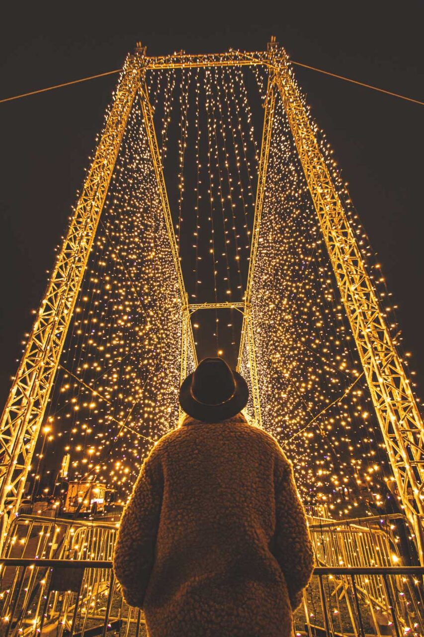 A person wearing a hat and coat looks at an illuminated light structure at a Christmas market in Europe.