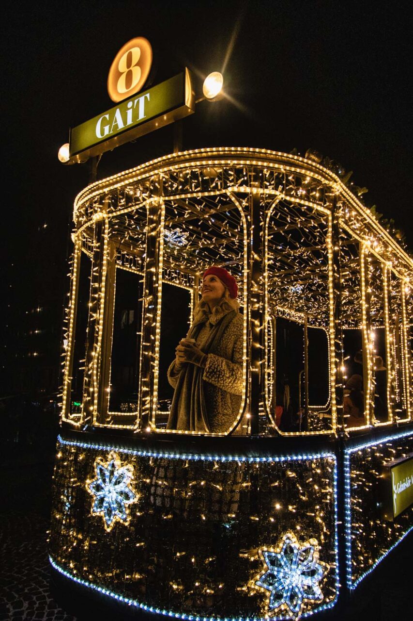 A woman in a red hat and teddy coat stands in a lit Christmas tram decorated with lights
