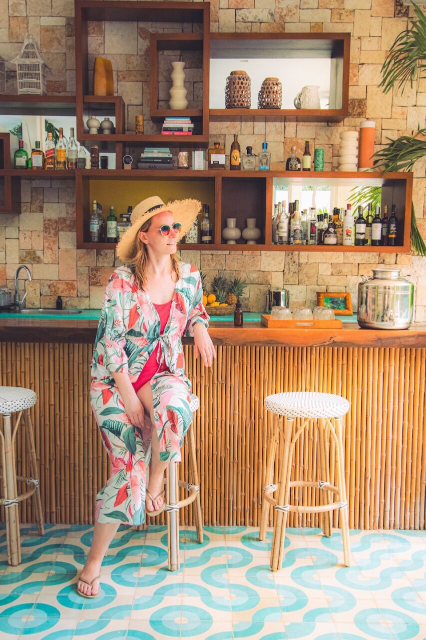 A woman in a hat and colorful swimsuit sits at a bamboo bar in Tiki Tiki Tulum - surrounded by tropical retro vibes, bottles gleaming behind her, and exotic details that feel straight out of a vintage postcard.