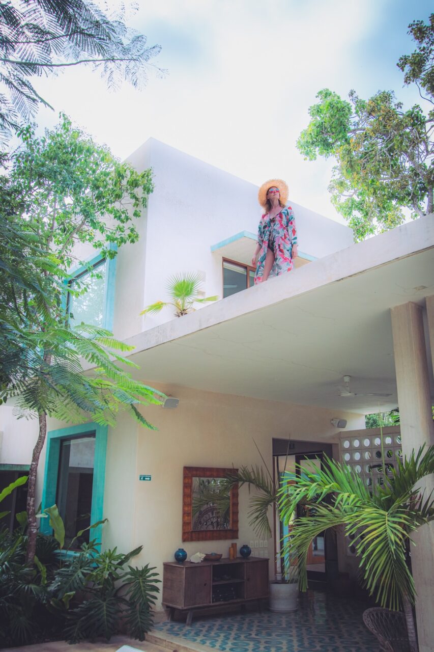 A woman in a straw hat stands on the terrace of Hotel Tiki Tiki Tulum, surrounded by lush tropical greenery, overlooking a serene patio with mirrors and jungle plants.