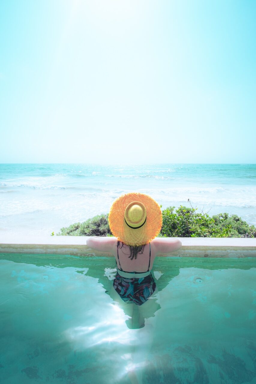 A woman in a straw hat sits in an infinity pool overlooking the ocean at Our Habitas Tulum. Tropical greenery and gentle waves in the background.