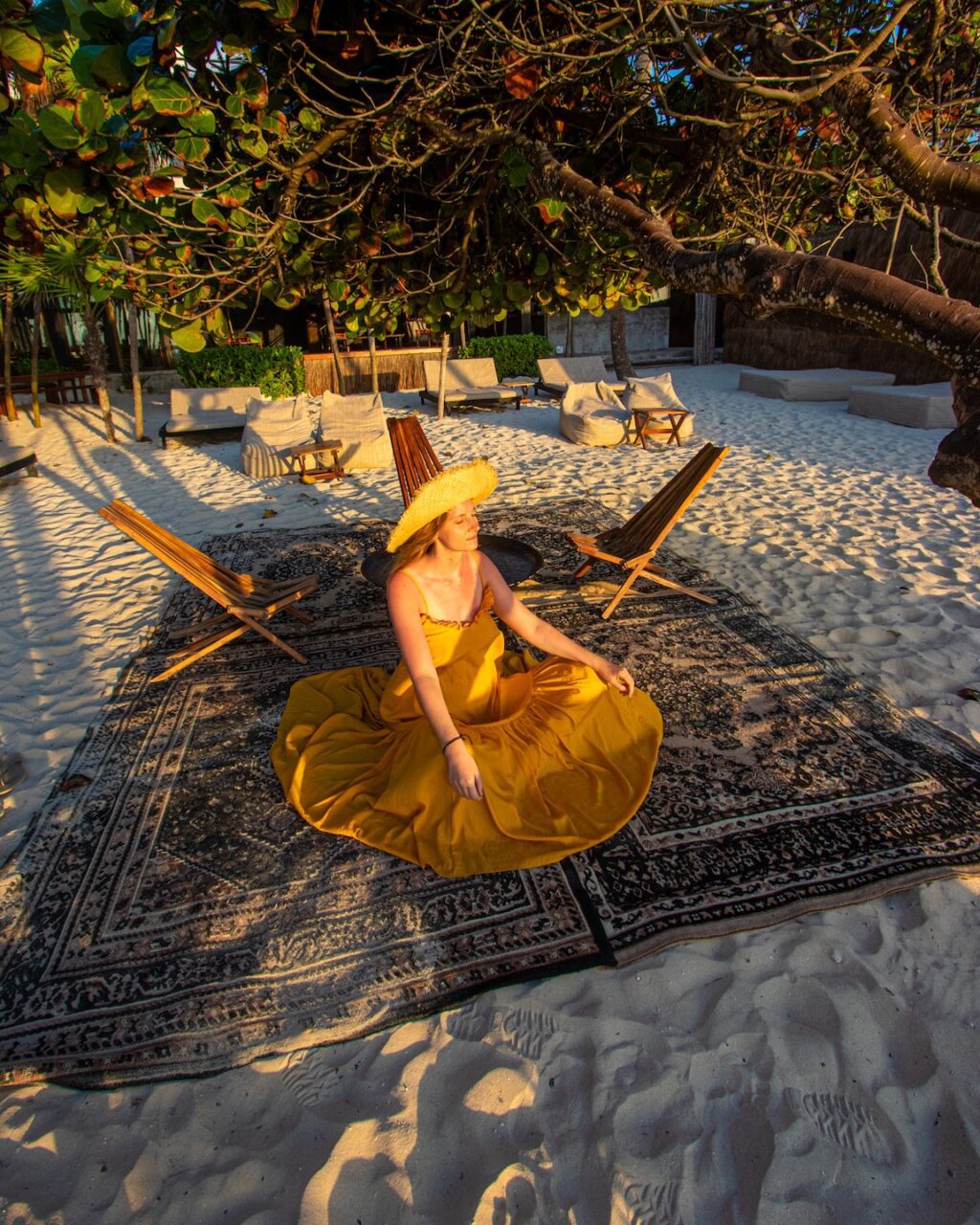 A woman in a yellow dress and straw hat sits on an oriental rug spread out on the beach in Tulum, under the shade of a tree, bathed in the warm light of the morning.