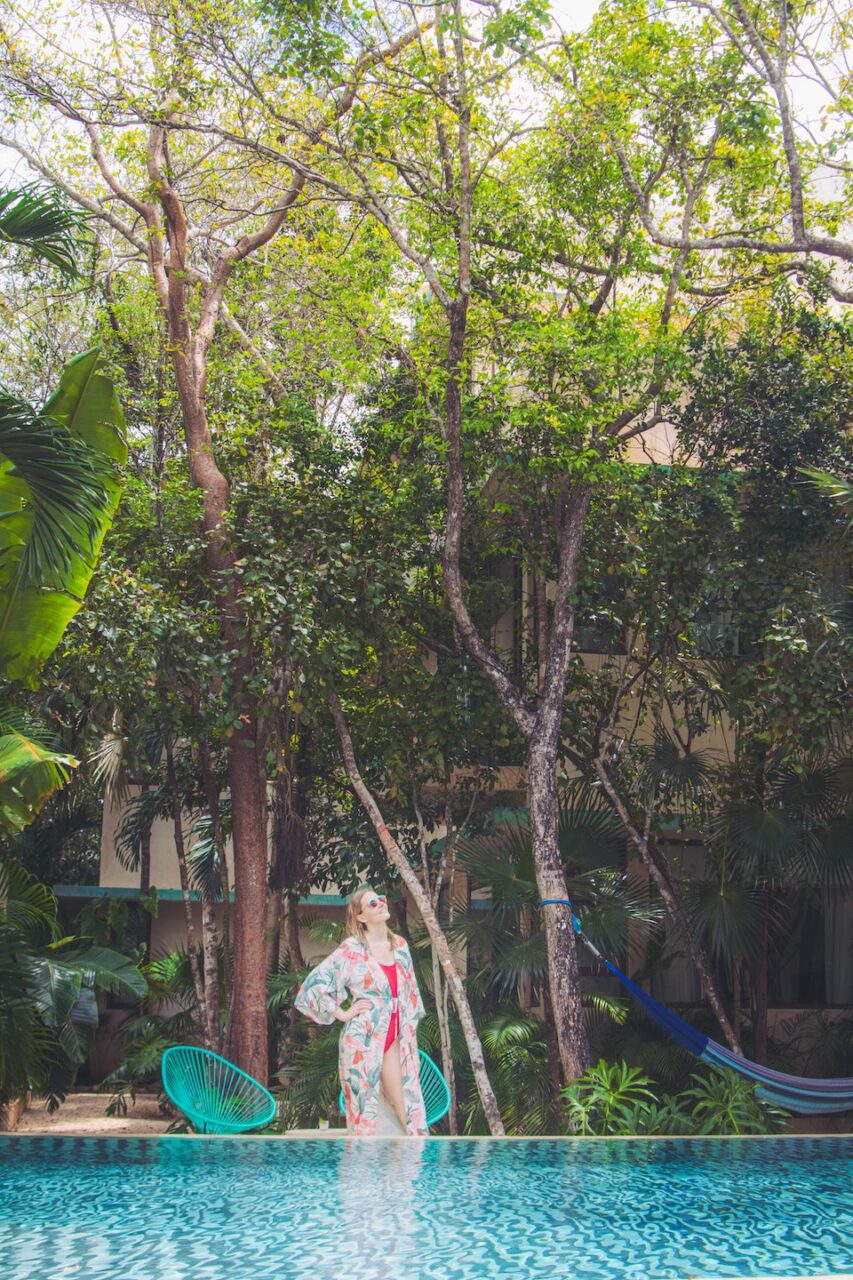 A woman in a colorful kimono and sunglasses stands by the intimate jungle pool at Tiki Tiki Tulum, surrounded by tropical greenery, a hammock, and turquoise chairs.