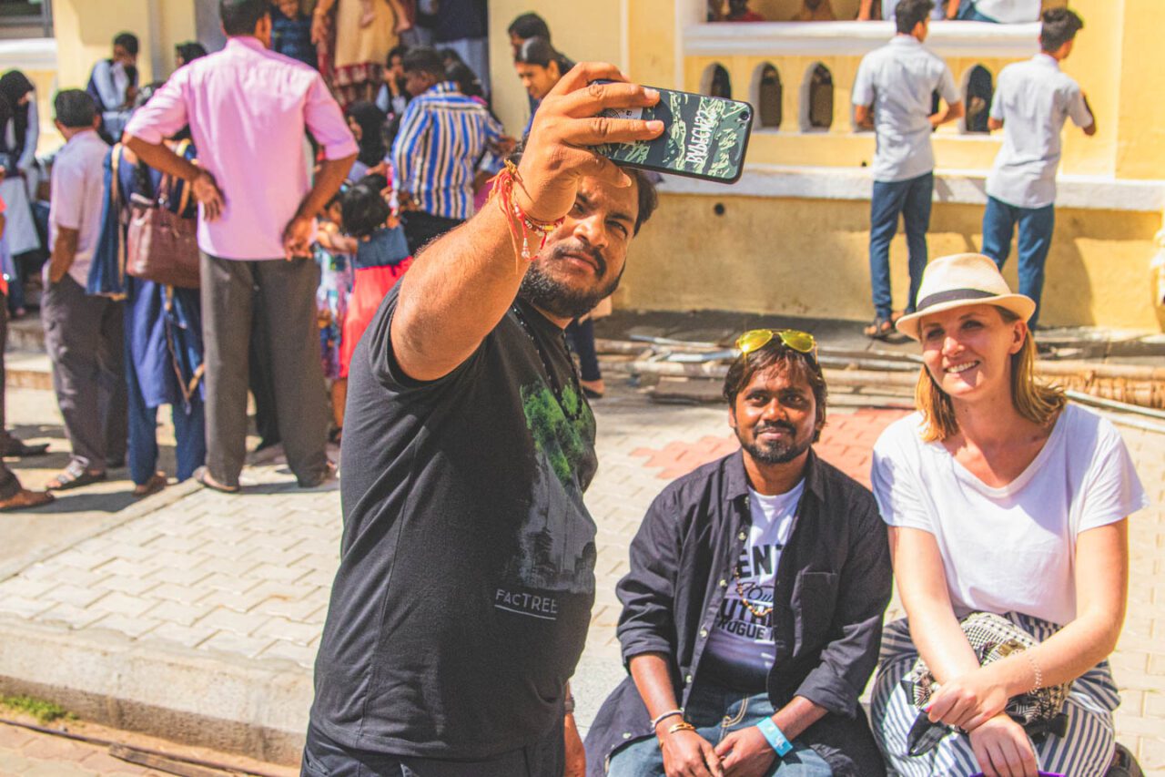 Grupa osób robiąca wspólne selfie w Mysore – kobieta w kapeluszu i dwóch mężczyzn uśmiechają się do aparatu, w tle tłum zwiedzających pałac.