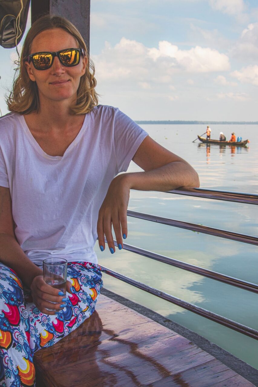 Kobieta w okularach przeciwsłonecznych siedząca na pokładzie houseboatu w Alleppey, Kerala – z relaksem w oczach i widokiem na spokojne wody i tradycyjną łódź w oddali.