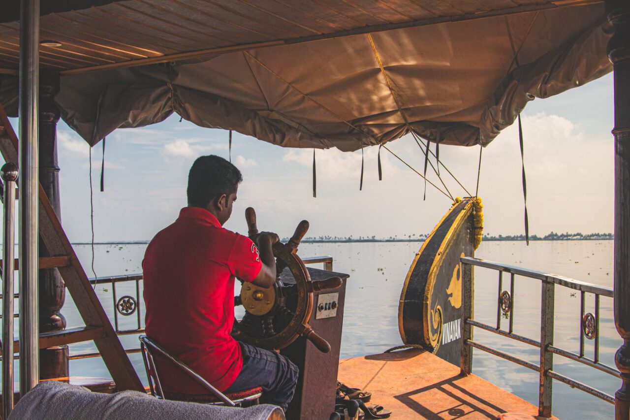 Mężczyzna w czerwonej koszulce steruje tradycyjną łodzią houseboat z otwartego pokładu.