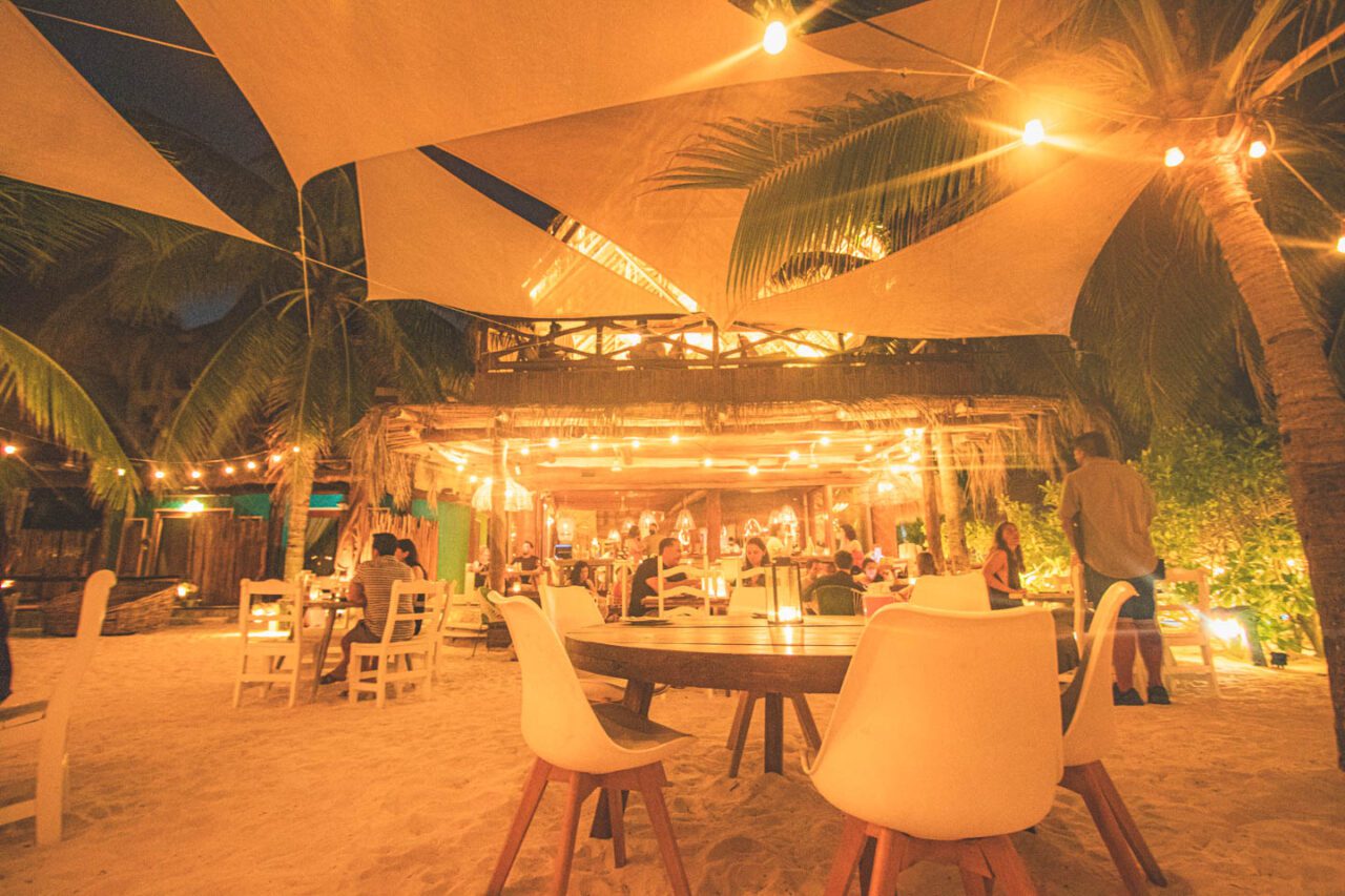 Wieczorna sceneria restauracji na plaży – Mandarina na Isla Holbox. Drewniane stoły i białe krzesła ustawione na piasku pod palmami i płóciennymi żaglami. Ciepłe światło żarówek oplatających palmy tworzy intymny, tropikalny klimat.