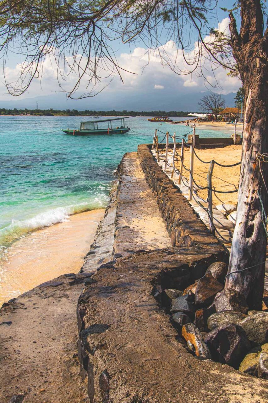 Tropical shoreline of Gili Trawangan with turquoise water, a small boat resting by the shore, a sandy beach and a stone promenade shaded by trees.
