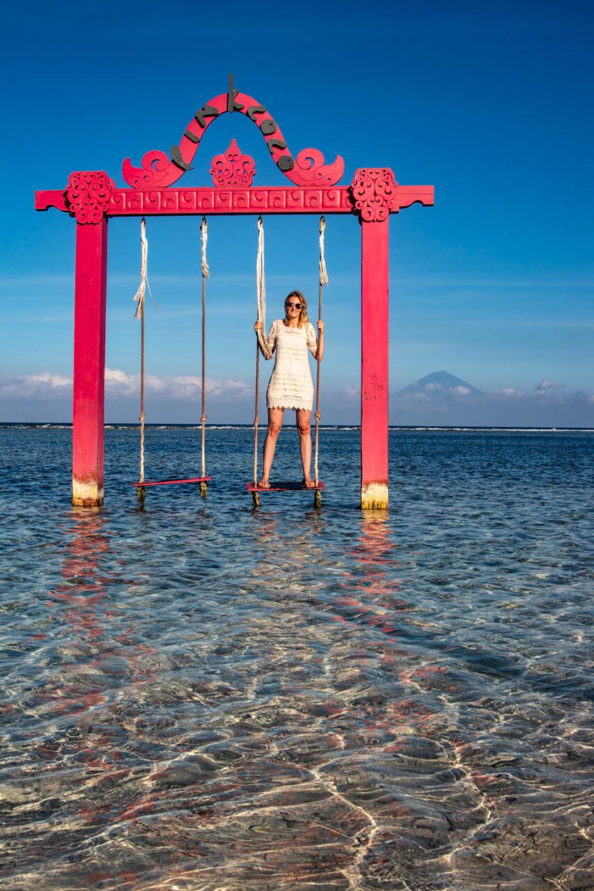 Woman standing on a red swing in the shallow, crystal-clear water of Gili Trawangan, with turquoise sea and Mount Agung visible in the background under a bright blue sky.