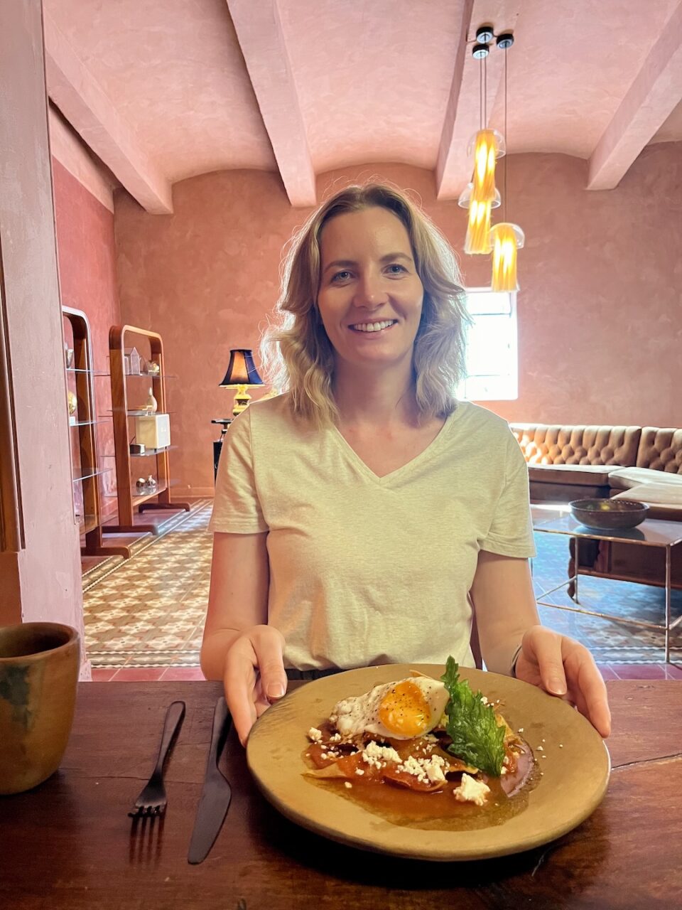 Breakfast at a hotel in Oaxaca, a woman sitting at a table and holding a plate with an egg and a local dish, the interior is in warm, pink tones.