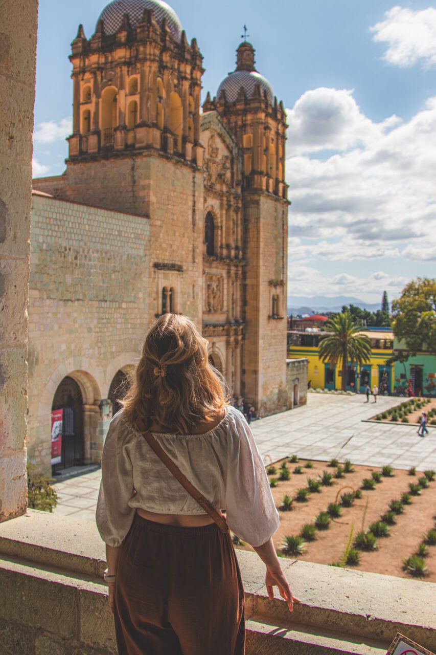 A view of the historic Santo Domingo de Guzmán church in Oaxaca, with a woman standing on a terrace and looking out at the historic city center.