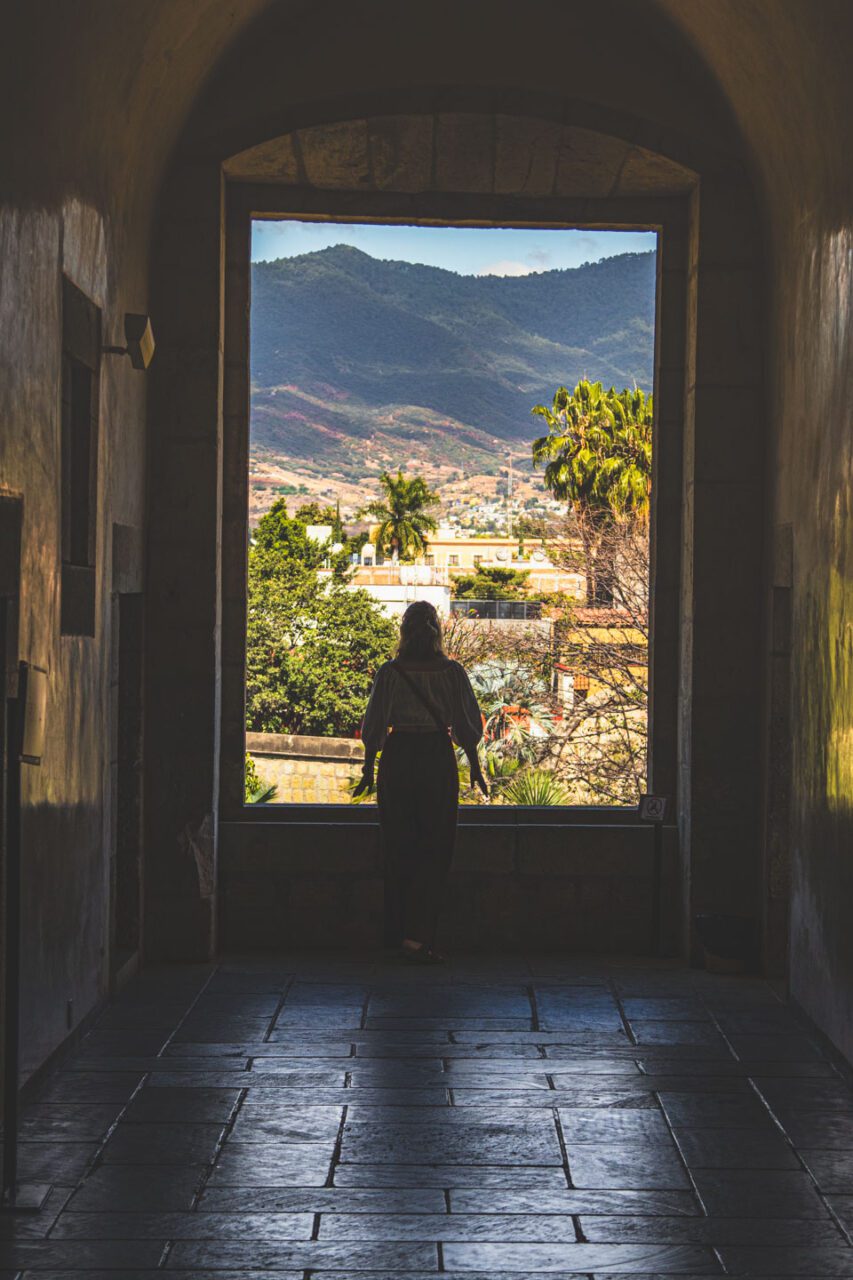 Kobieta stojąca w kamiennym przejściu dawnego klasztoru Santo Domingo w Oaxaca, patrząca na zieloną dolinę i góry w oddali.