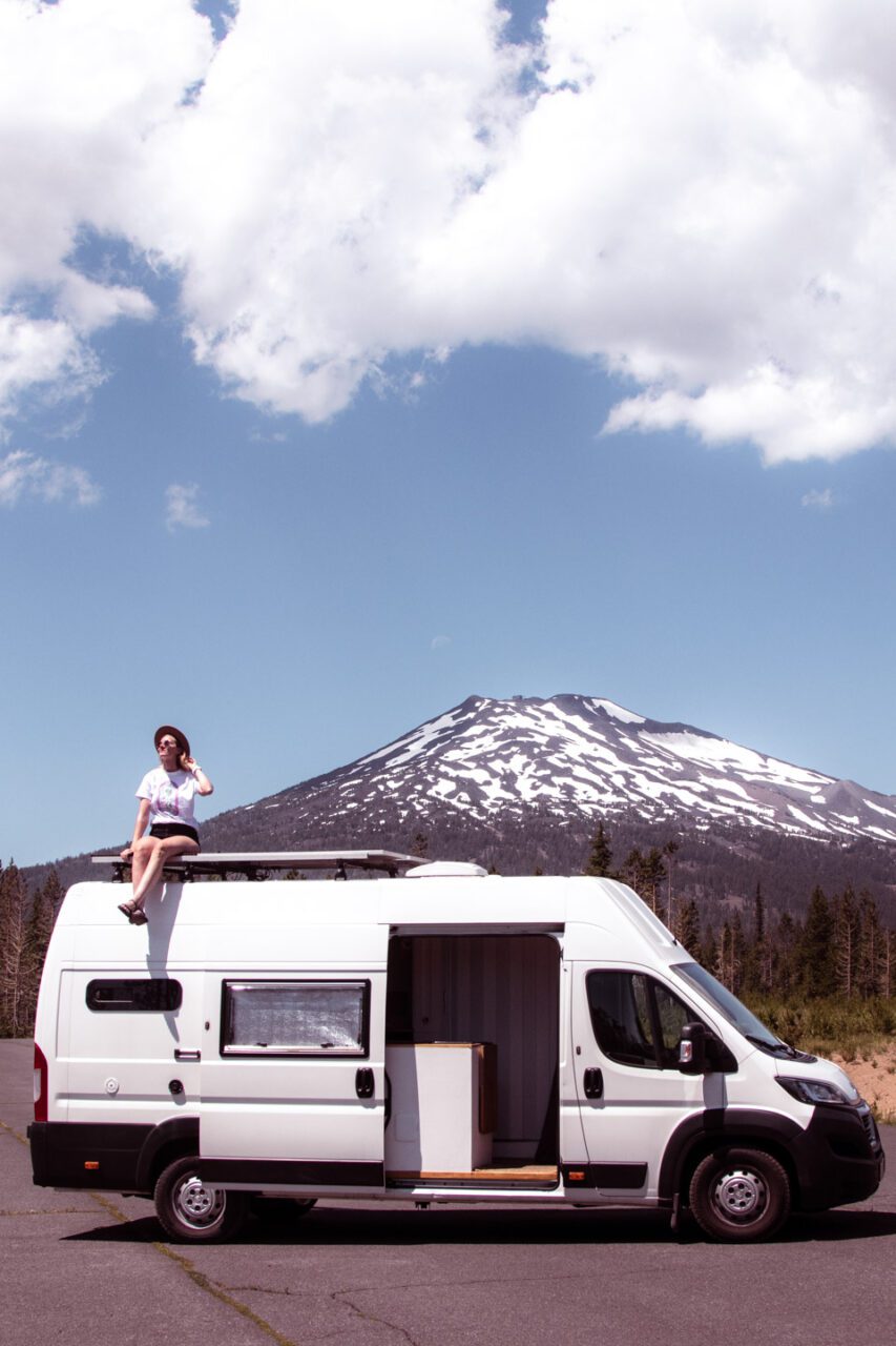 A camper van with a woman on the roof, set against the backdrop of a volcano near Bend, Oregon.