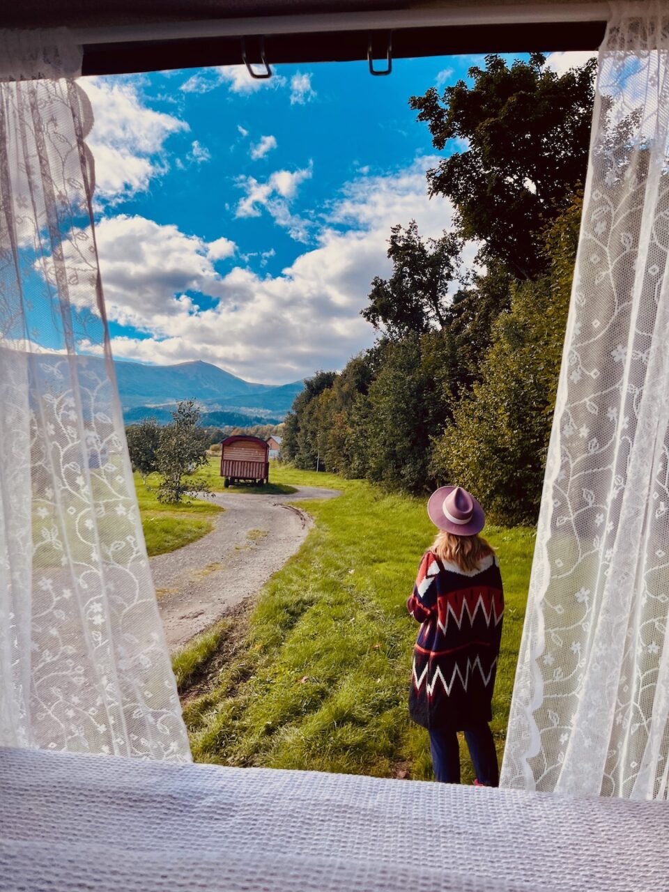 Widok z kampera przez koronkową firankę na kobietę w kapeluszu patrzącą na górską ścieżkę