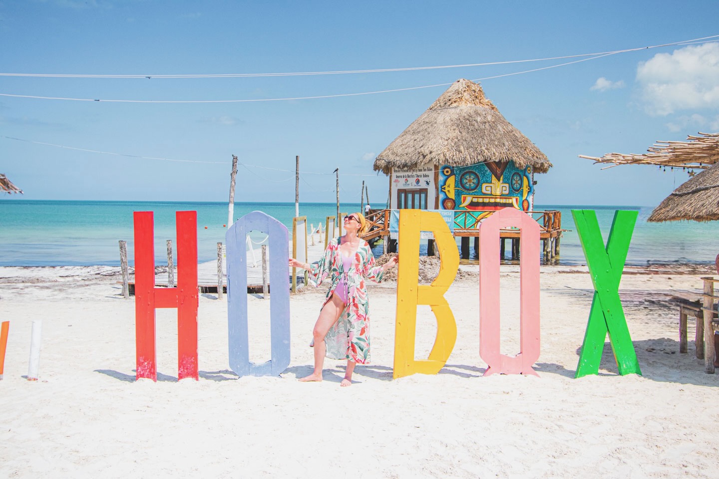 Woman standing barefoot on the beach in front of colorful HOLBOX sign, with turquoise ocean and palapa huts in the background – sunny island vibes.
