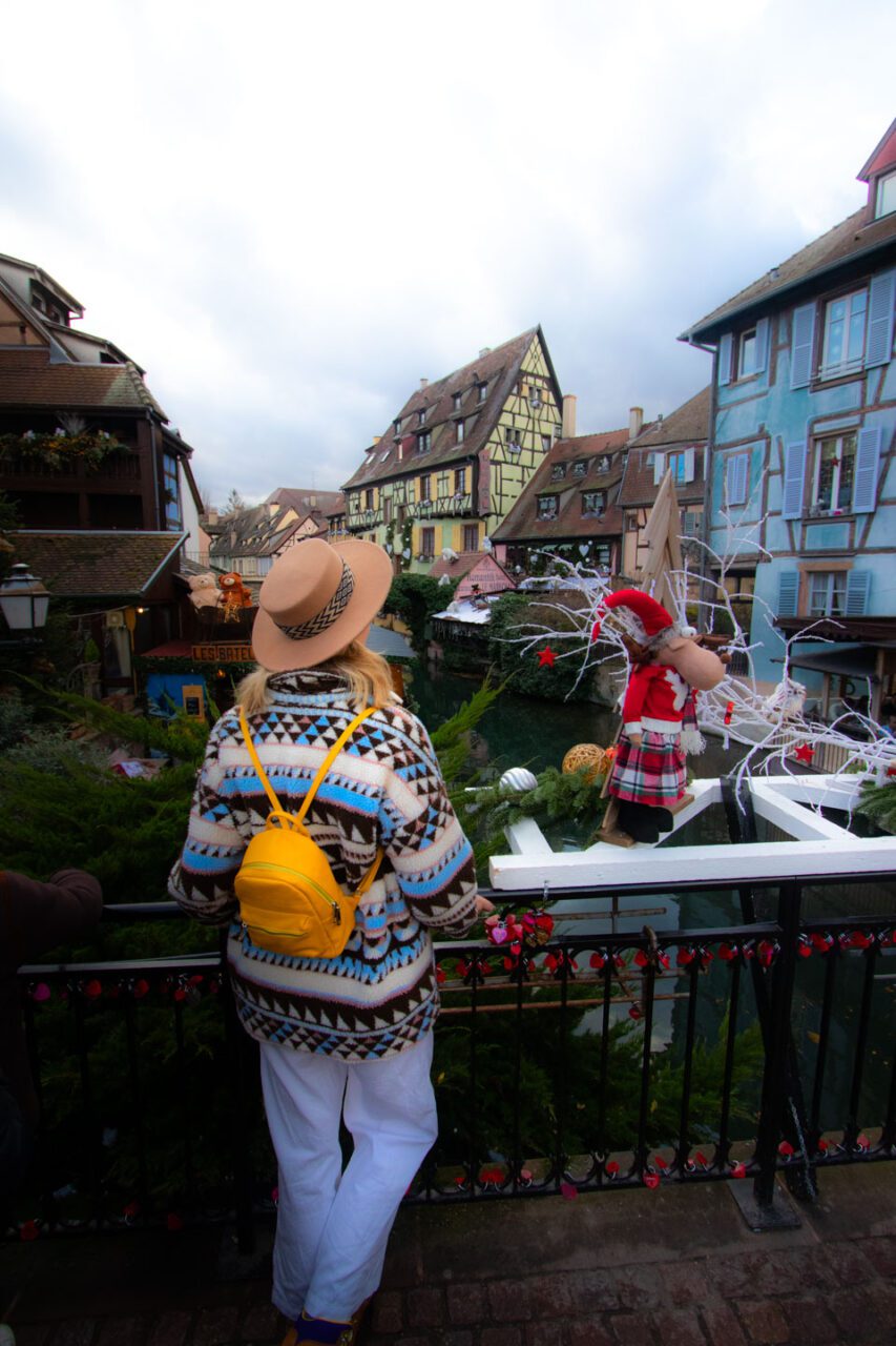 A woman in a hat and yellow backpack looks out at the canal and colorful half-timbered houses in festive Colmar.