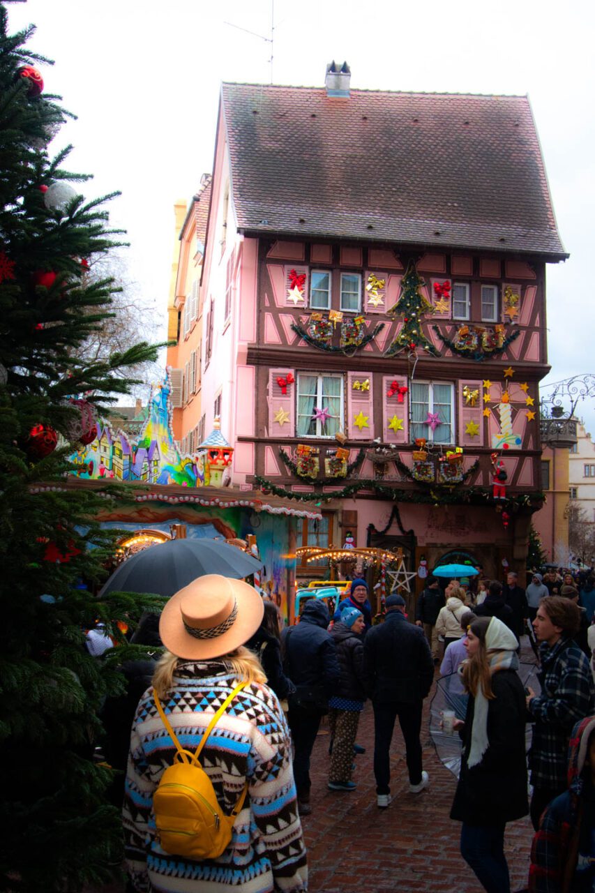 A tourist wearing a colorful sweater and straw hat looks at a pink house decorated for the holidays in Colmar during the Christmas market