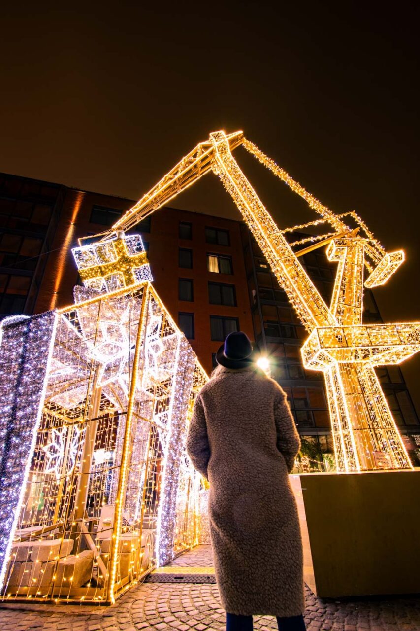 A woman in a teddy coat and hat looks at a shipyard crane illuminated by a lighting installation at the Christmas market in Gdańsk