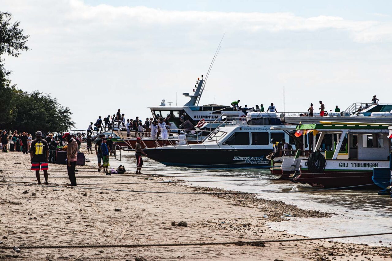 Zatłoczony port na Gili Trawangan z turystami wsiadającymi do łodzi i walizkami leżącymi na plaży.