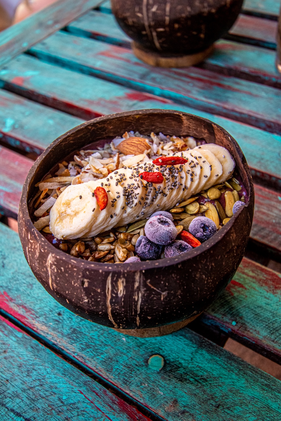 Kolorowa miska acai z plasterkami banana, mrożonymi jagodami, nasionami chia, pestkami i granolą, podana w kokosowej misce na turkusowym, przecieranym stole.