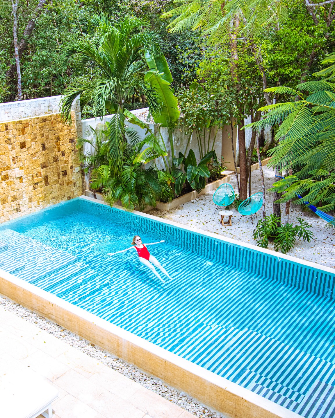 Kobieta w czerwonym stroju kąpielowym dryfuje na plecach w biało-turkusowym basenie otoczonym palmami w hotelu Tiki Tiki Tulum. Tropikalna sceneria, fotele Acapulco i artystyczna estetyka retro.