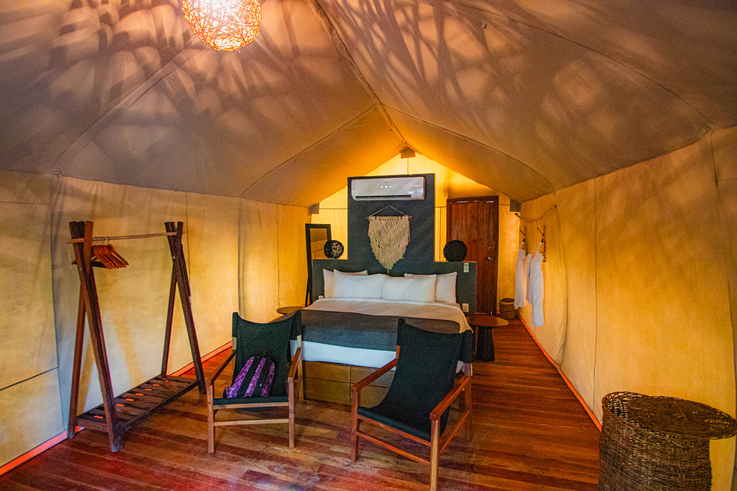 Wnętrze namiotu glampingowego z dużym łóżkiem, dwoma czarnymi fotelami, drewnianą podłogą i dekoracyjnym światłem w stylu boho, w hotelu Our Habitas Tulum.