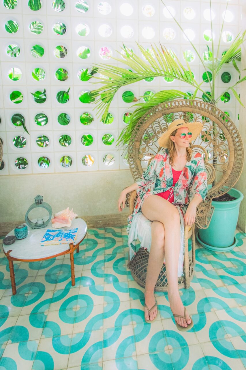 A woman in a swimsuit and sunhat relaxes in a decorative rattan chair at Tiki Tiki Tulum - a bright, tropical space with colorful tile floors, palm leaves, and a white wall dotted with round cutouts framing the jungle greenery outside.