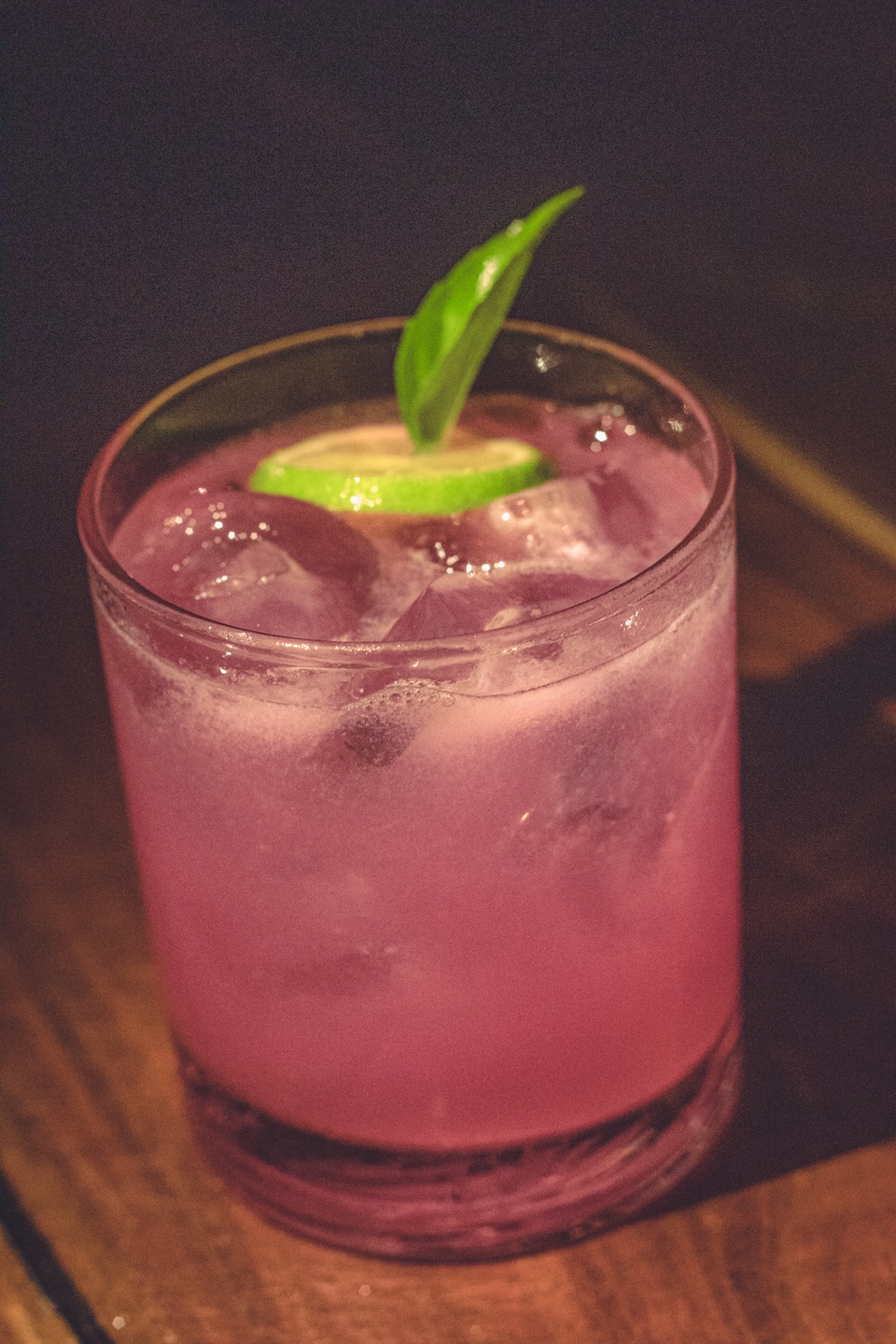 A pink hibiscus cocktail served over ice in a lowball glass, garnished with a slice of lime and a basil leaf, photographed in the evening on a wooden table at Our Habitas Tulum.