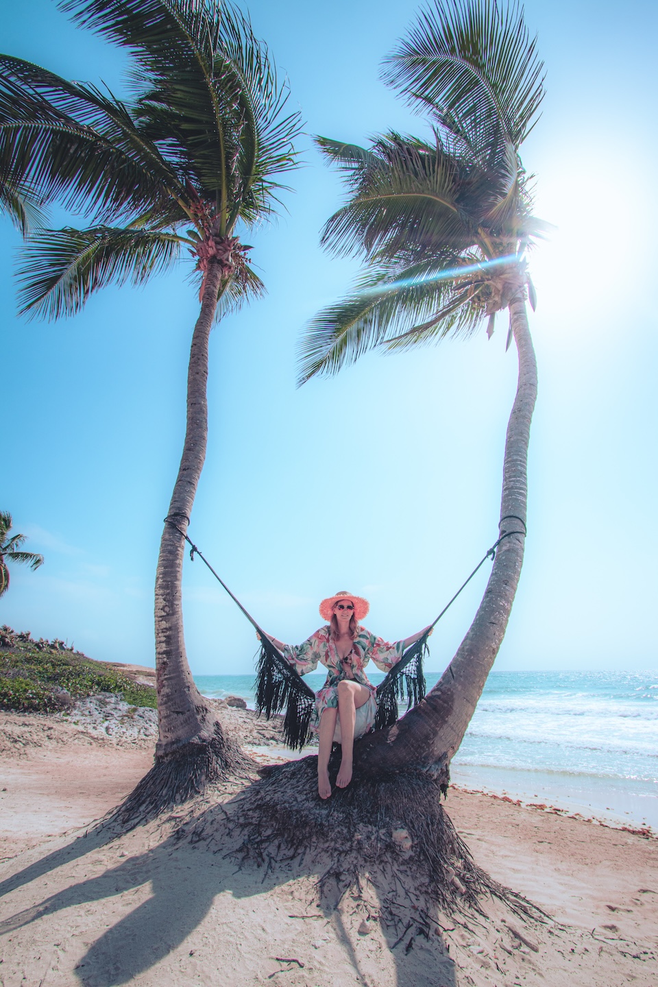 Woman in a tropical dress and straw hat relaxing in a hammock between two palm trees on a beach in Tulum, with the ocean and blue sky in the background.