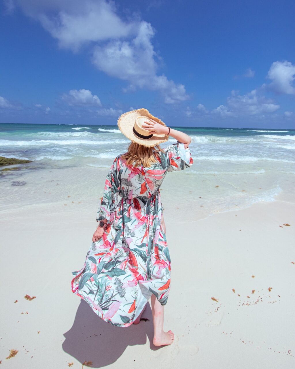 A woman in a colorful dress and straw hat walks barefoot along the white sand beach in Tulum, gazing toward the turquoise sea and a sky dotted with white clouds.