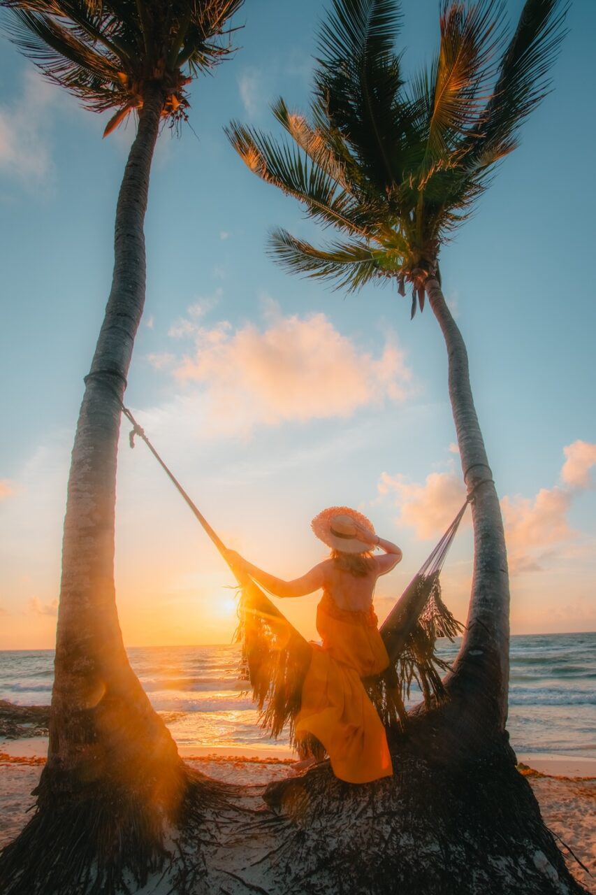 A woman in an orange dress relaxes in a beach hammock at sunrise in Tulum, framed by palm trees and soft morning light.