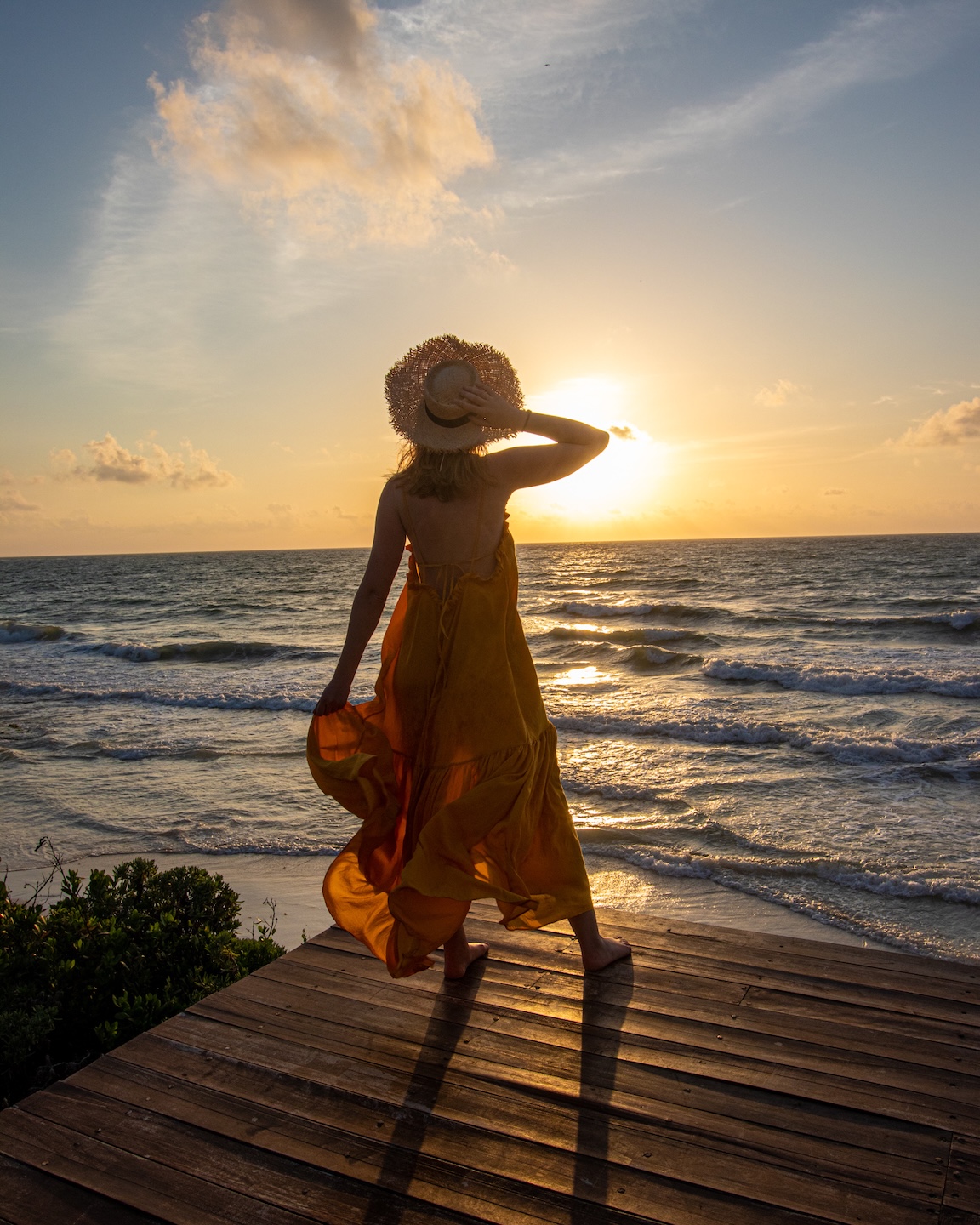 A woman in a flowing yellow dress and straw hat watches the sunrise over the ocean in Tulum, standing barefoot on a wooden deck by the beach.
