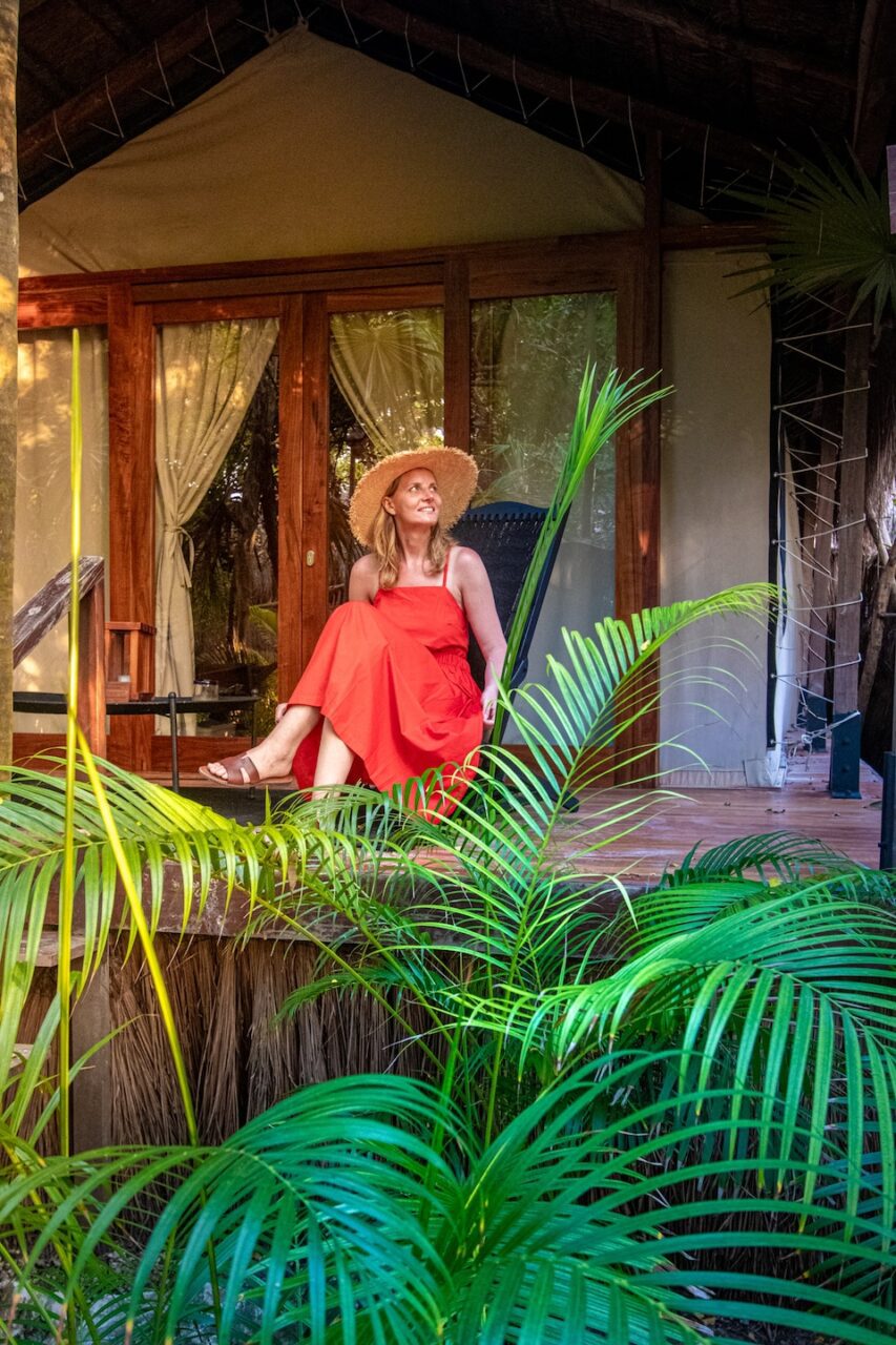 A woman in a red dress and straw hat sits on the veranda of a glamping tent surrounded by lush jungle palms in Tulum.