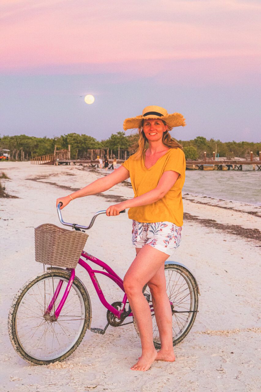 Kobieta w słomkowym kapeluszu prowadzi różowy rower po plaży na Isla Holbox o zachodzie słońca, w tle pełnia księżyca i pastelowe niebo.