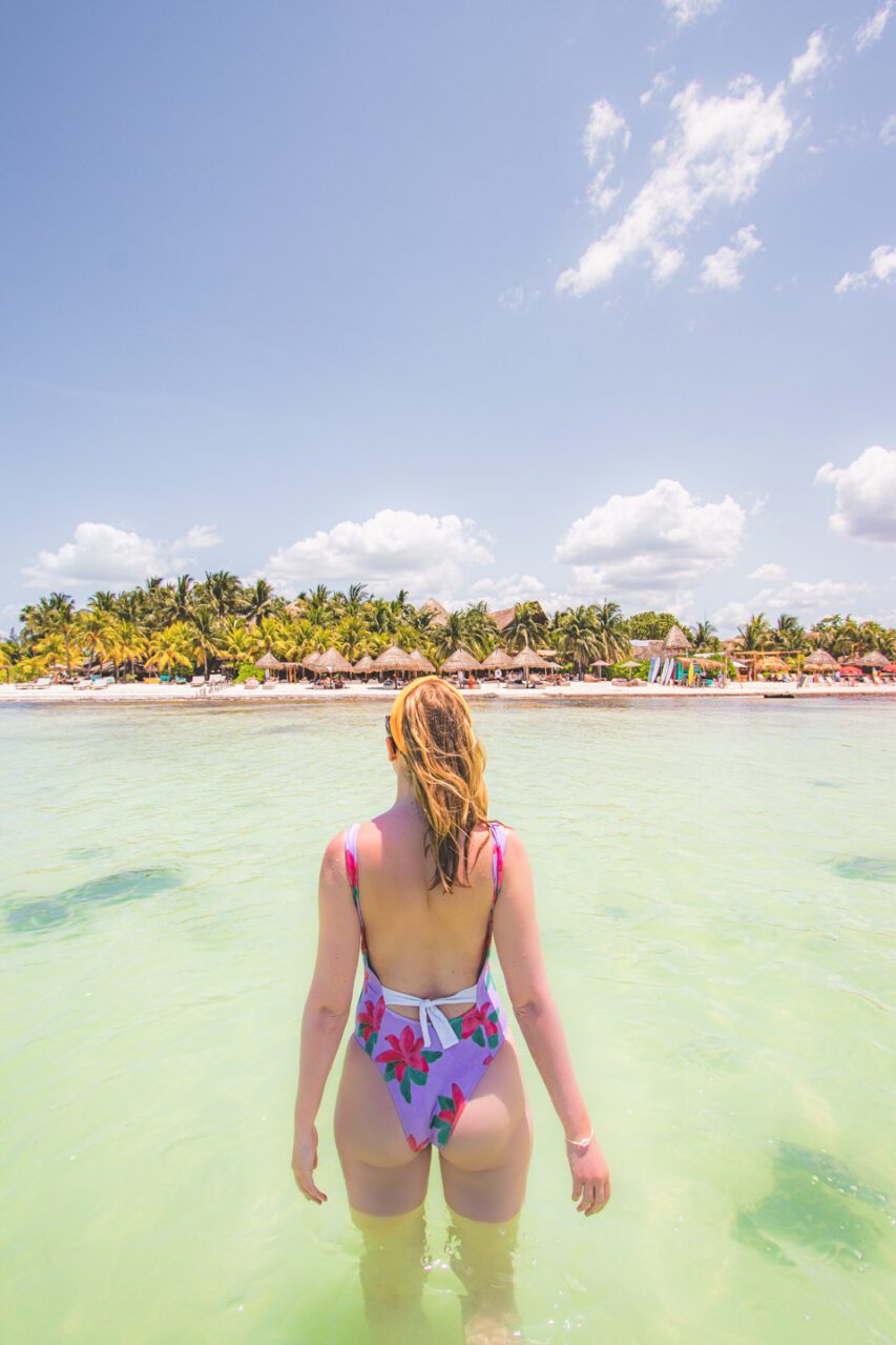 Kobieta w kolorowym stroju kąpielowym wchodzi do wody na Isla Holbox – tropikalna plaża i palmy w tle.