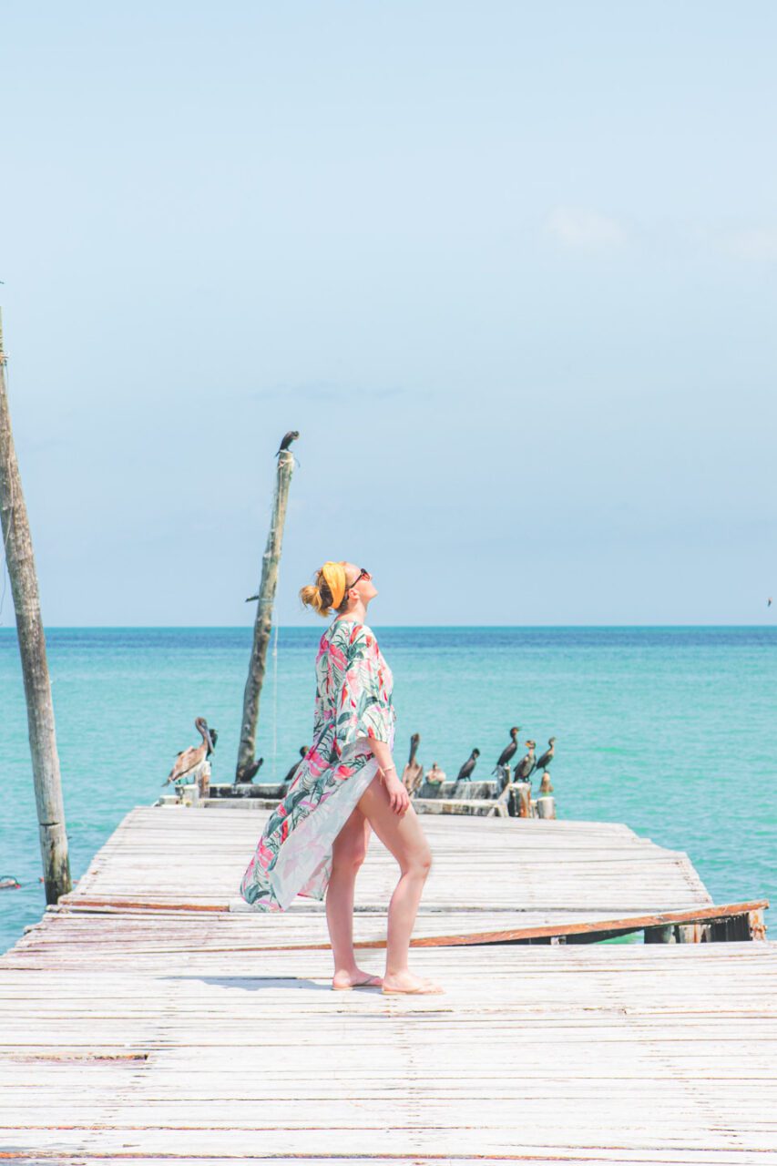 Kobieta w pareo spacerująca po drewnianym pomoście na Isla Holbox, otoczona turkusowym morzem i ptakami wodnymi – spokojny, słoneczny moment nad oceanem.