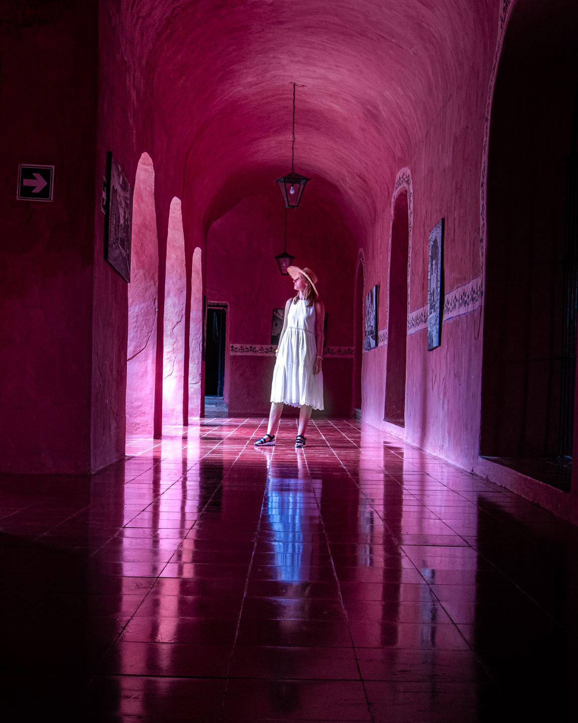 Kobieta w białej sukience stoi w różowym, sklepionym korytarzu z odbiciem światła na błyszczącej podłodze.