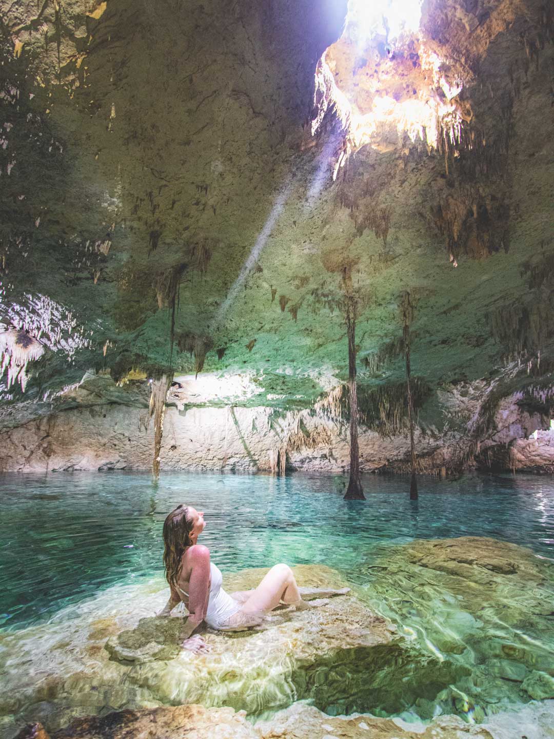 A woman sitting on a rock inside a cenote in Mexico, surrounded by clear turquoise water and illuminated by a beam of sunlight coming through an opening in the cave ceiling.