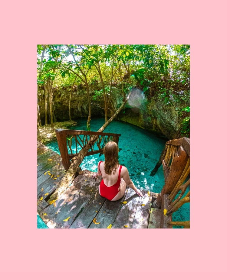A woman sitting by a cenote in Mexico, surrounded by turquoise water and jungle vegetation.