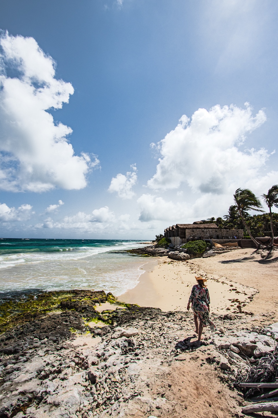 A woman wearing a hat walks alone on a wild beach in Tulum, overlooking waves, rocks, and rustic cabanas nestled among palm trees.