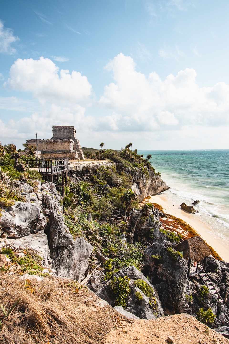 The Mayan ruins of Tulum are situated on a cliff overlooking the Caribbean Sea, surrounded by vegetation, with a beach access below and a blue sky in the background.