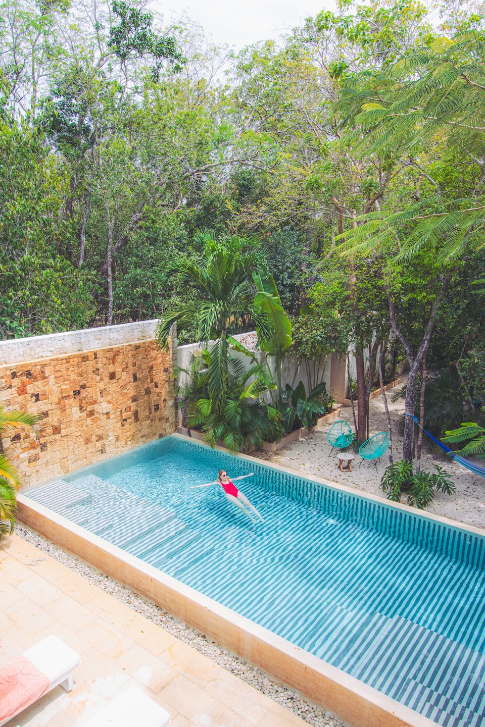A woman in a red swimsuit floats on her back in a geometric pool at the Tiki Tiki hotel in Tulum, surrounded by tropical vegetation and palm trees.