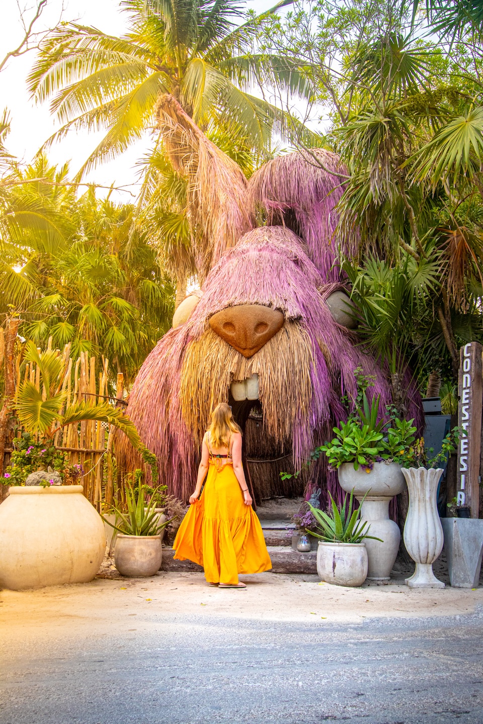 A woman in a yellow dress stands in front of the fairytale entrance to the Conestesia restaurant in Tulum, Mexico – an iconic spot for Instagram photos surrounded by palm trees and exotic vegetation.