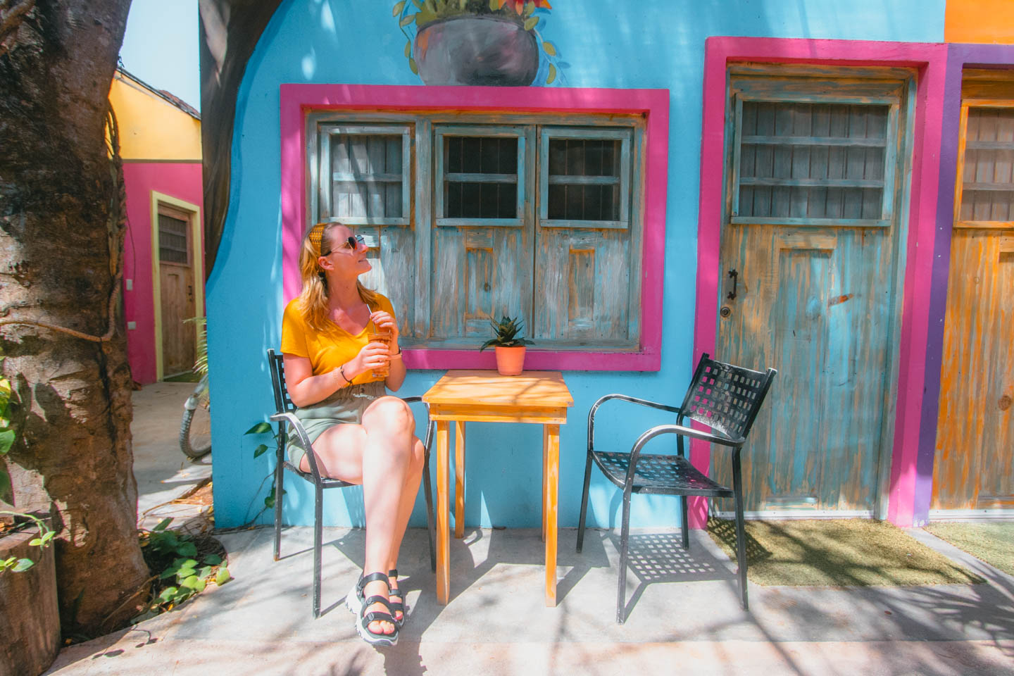 A woman in a yellow blouse and sunglasses sits at a colorful table in front of a turquoise and pink wall in Tulum, Mexico.