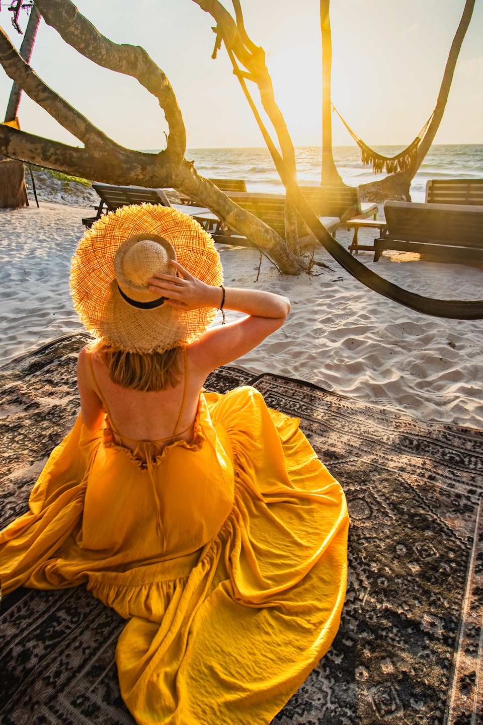 A woman in a yellow dress and straw hat sits on a beach in Tulum at sunrise, surrounded by hammocks and trees