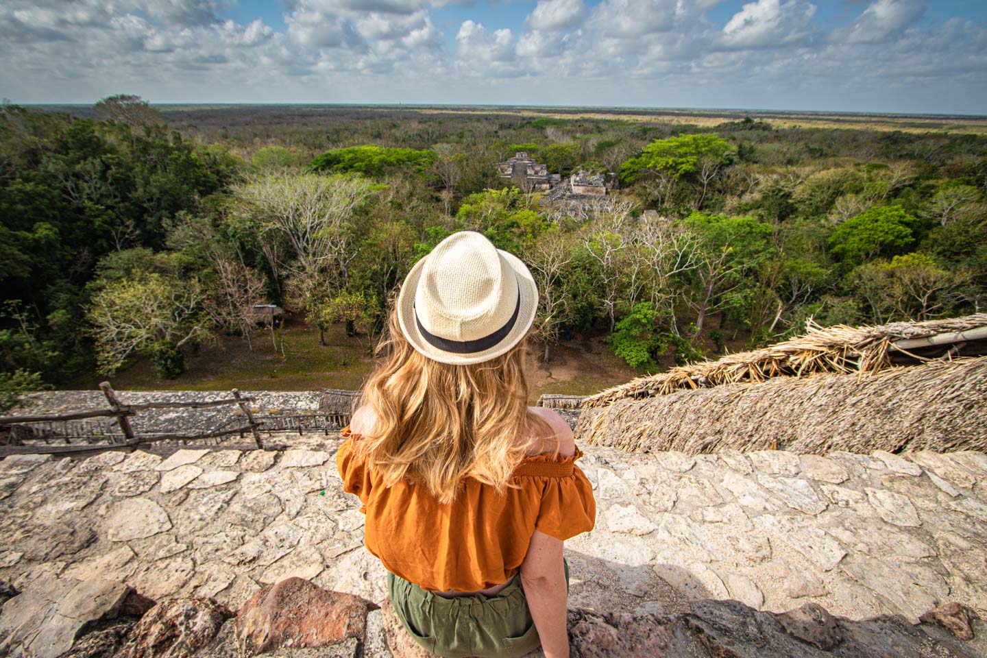 A woman wearing a hat sits atop a Mayan pyramid, looking out over the jungle and ruins of Ek Balam in Yucatan, Mexico