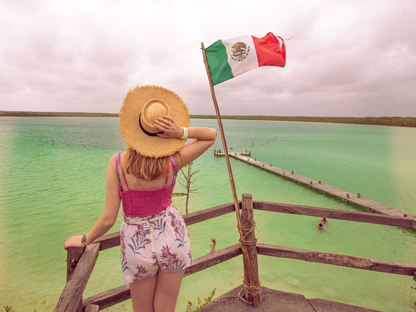A woman wearing a hat looks out over the Kaan Luum lagoon in Tulum, Mexico – the view from a platform with a Mexican flag and a jetty over the turquoise water