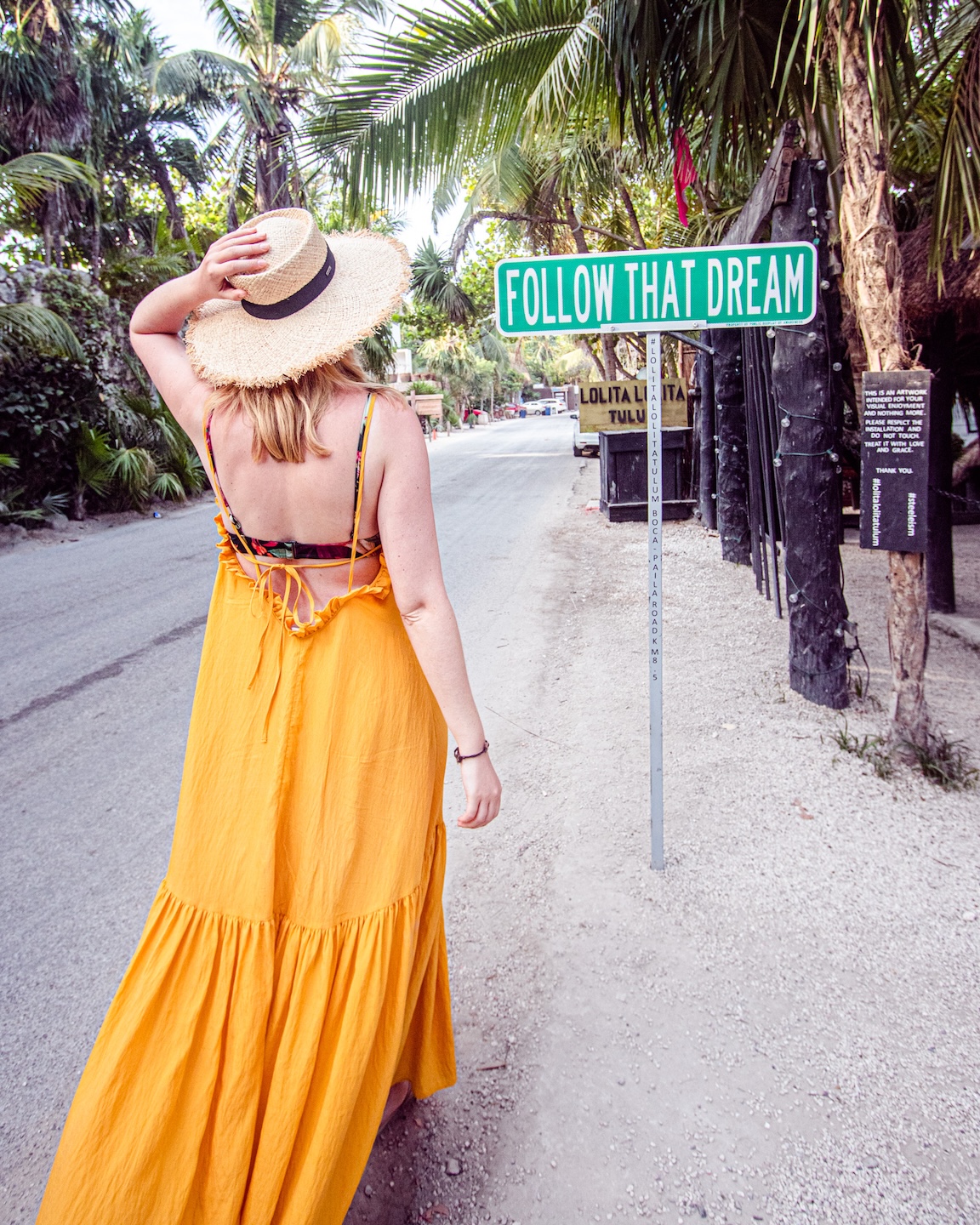 A woman in a yellow dress and hat walks toward a green "Follow That Dream" sign on a palm-lined street in Tulum, Mexico.